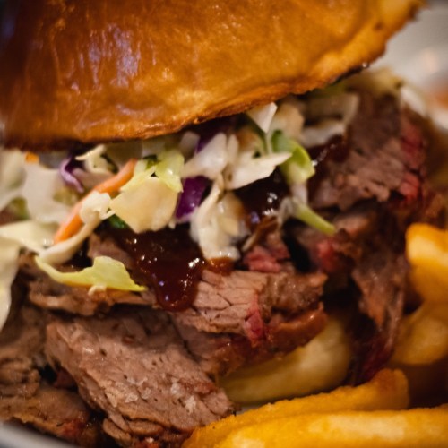 Close-up of a hearty sandwich with sliced beef, coleslaw, and barbecue sauce, served with crispy fries.