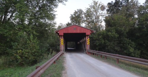 A red covered bridge surrounded by greenery, with a gravel road leading to it and warning signs on either side.