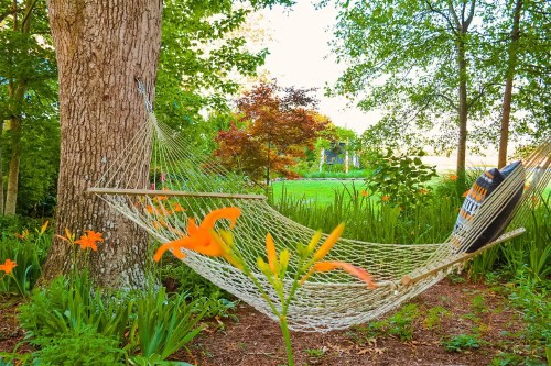 A hammock hangs between two trees, surrounded by vibrant flowers and lush greenery in a serene garden setting.