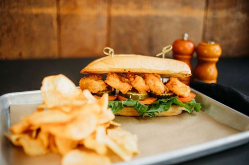 A crispy fried shrimp sandwich with lettuce, tomato, and pickles, served with a side of potato chips.