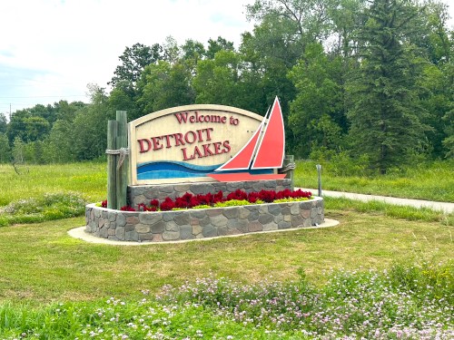 Welcome sign for Detroit Lakes, featuring a sailboat design and surrounded by colorful flowers and greenery.