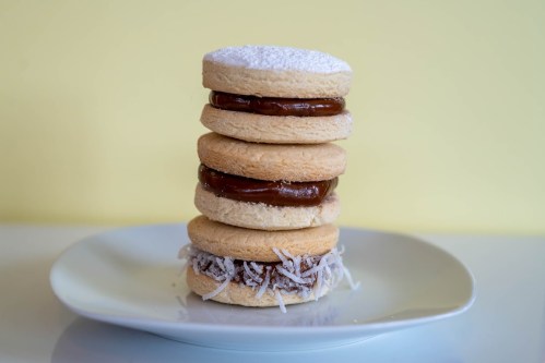 A stack of four sandwich cookies with caramel filling, topped with powdered sugar and coconut flakes on the bottom cookie.