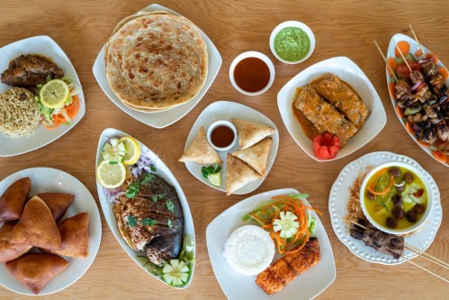 A variety of colorful dishes including fried fish, kebabs, samosas, and flatbreads arranged on white plates.