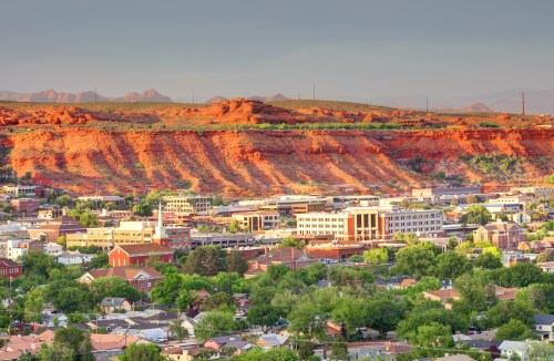 A scenic view of a town with green trees and buildings, set against striking red rock formations in the background.
