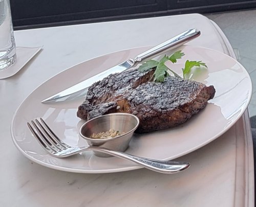 A grilled steak on a white plate, garnished with parsley, accompanied by a small bowl of seasoning and cutlery.