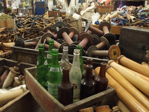 A cluttered antique shop filled with wooden spools and vintage bottles in a wooden crate.