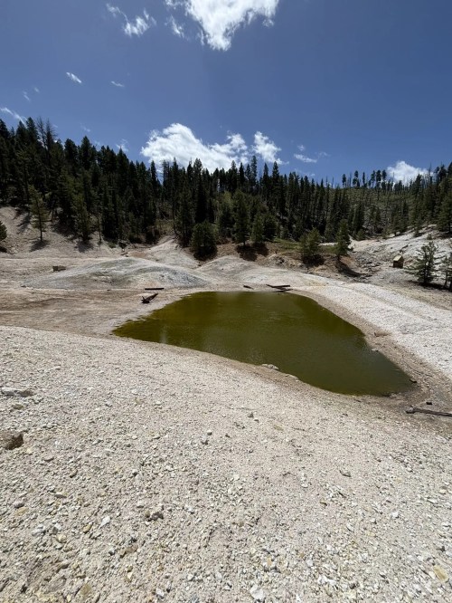 A small, greenish pond surrounded by dry, rocky terrain and sparse trees under a blue sky with scattered clouds.