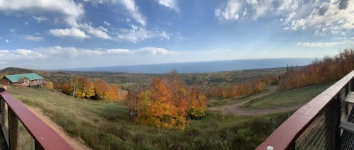 Panoramic view of a colorful autumn landscape with trees and a lake in the distance under a partly cloudy sky.