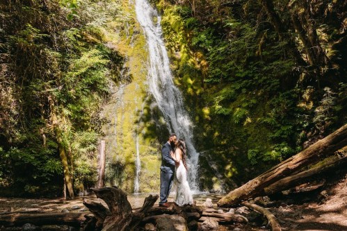 A couple embraces in front of a lush waterfall, surrounded by greenery and fallen logs in a serene forest setting.