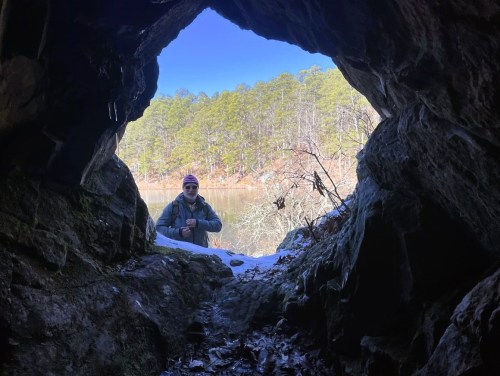 A person stands in a cave entrance, looking out at a scenic view of trees and water on a clear day.