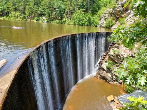 A scenic view of a waterfall cascading over a curved dam into a calm lake, surrounded by lush greenery and rocks.
