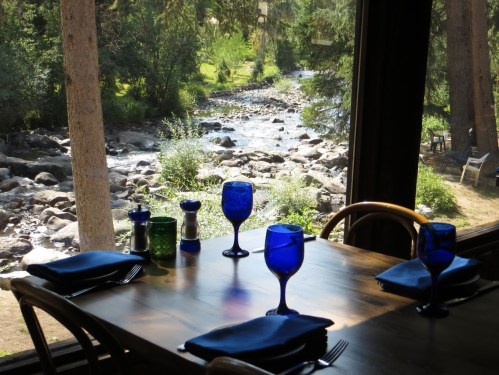 A dining table set with blue glasses overlooks a serene river and lush greenery through a large window.