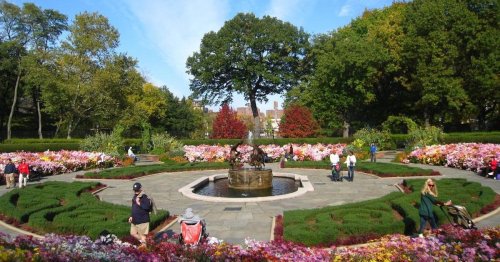 A vibrant garden with a central fountain, surrounded by colorful flowers and people enjoying the scenery on a sunny day.