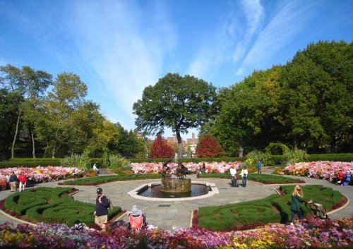A vibrant garden with colorful flowers, a central fountain, and people enjoying the scenery under a blue sky.