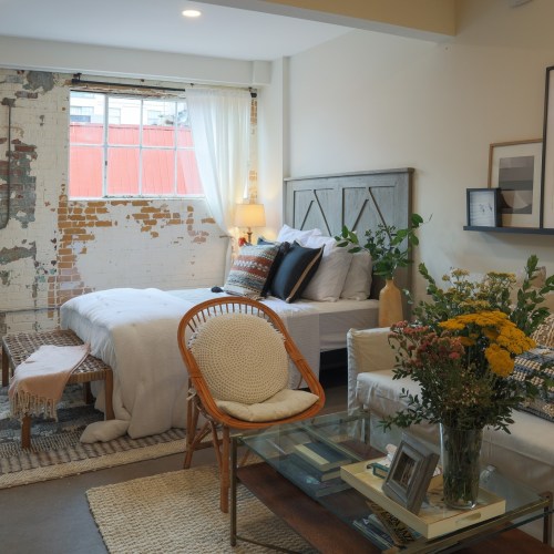 Cozy bedroom with a bed, plants, and a seating area, featuring exposed brick walls and natural light.