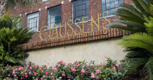Sign reading "CLAUSSEN'S" on a brick building, surrounded by lush greenery and colorful flowers.