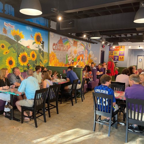 A vibrant restaurant scene with diners at tables, colorful murals, and a lively atmosphere filled with people.