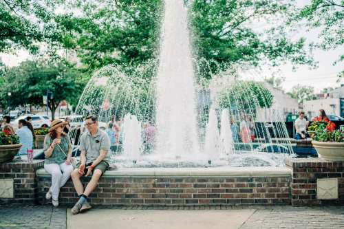 A couple sits by a fountain in a park, surrounded by flowers and people enjoying the sunny day.