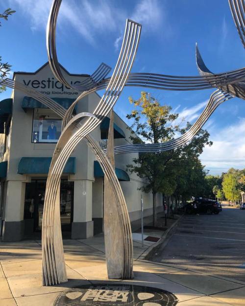 A modern metal sculpture stands in front of a boutique, with trees and a blue sky in the background.