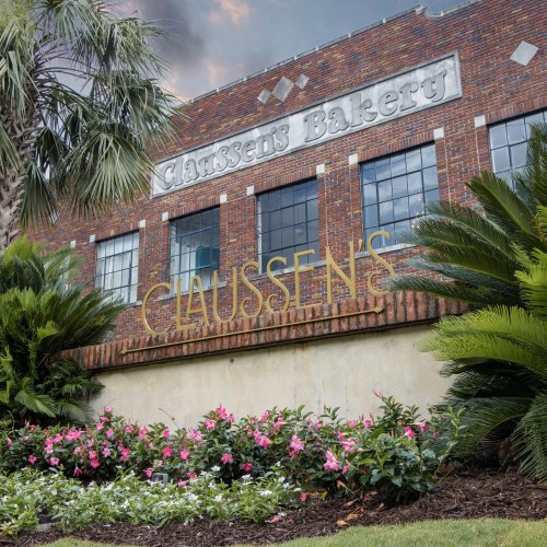 A brick building with "Claussen's Bakery" sign, surrounded by lush greenery and colorful flowers.