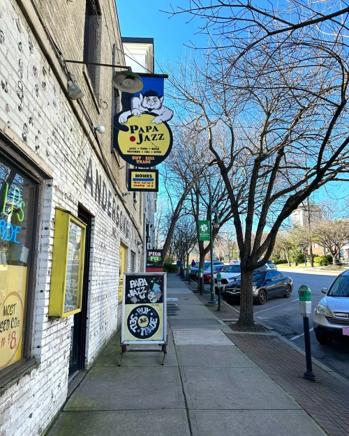 A sidewalk view of a street with trees, featuring a sign for "Papa Jazz" and nearby storefronts.