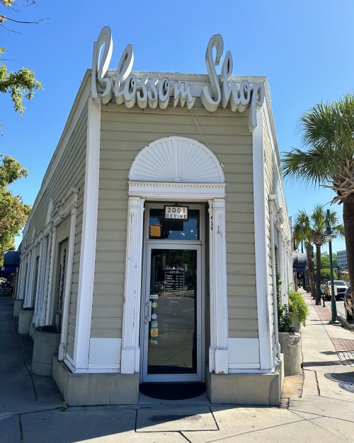 Exterior of the Blossom Shop, a light-colored building with a decorative entrance and palm trees nearby.