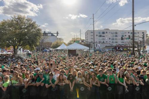 A large crowd in green attire celebrates outdoors under a sunny sky, with tents and buildings in the background.