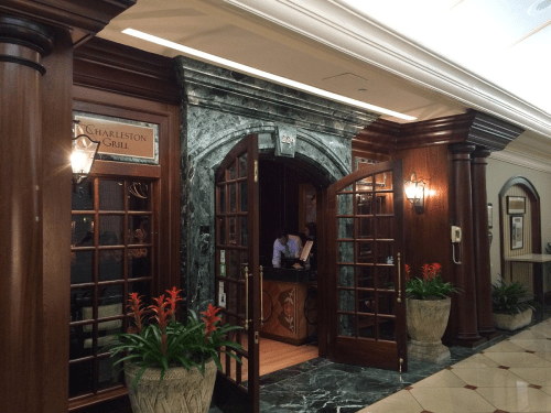 Entrance to Charleston Grill featuring wooden doors, marble accents, and potted plants. Warm, inviting atmosphere.