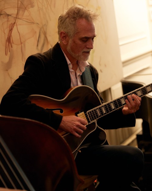 A musician playing an acoustic guitar, focused and seated, with a soft background of abstract art.