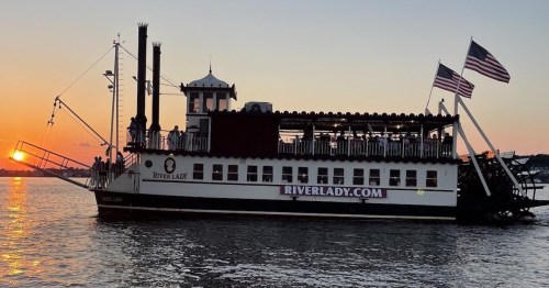 A paddleboat named "River Lady" sails on water at sunset, with American flags waving and people on the upper deck.