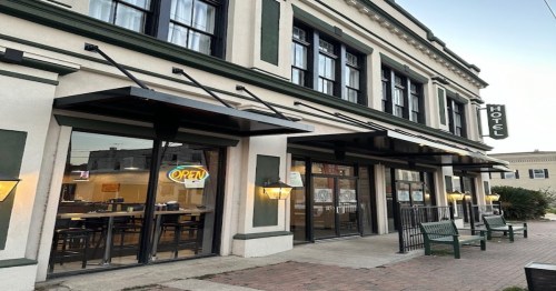 Exterior of a hotel with large windows, an "Open" sign, and outdoor seating. The building features classic architecture.