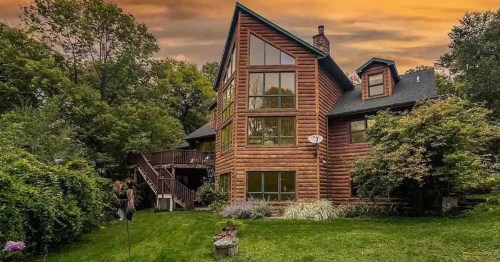 A large log cabin with multiple windows, surrounded by greenery and a sunset sky in the background.