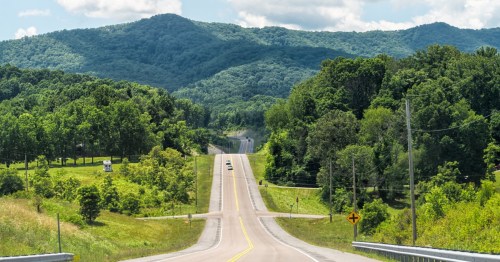 A winding road leads through lush green hills under a bright blue sky with scattered clouds.