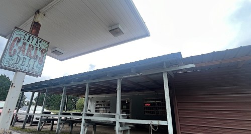 A rustic deli with a weathered sign, outdoor seating, and a partially cloudy sky overhead.