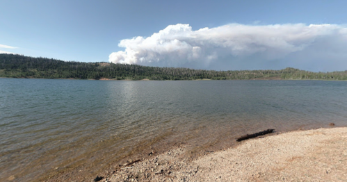 A serene lake with a rocky shore, surrounded by trees, and a large cloud of smoke rising in the distance.