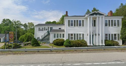 A white two-story building with columns, surrounded by greenery, along a road with a sign visible in the foreground.