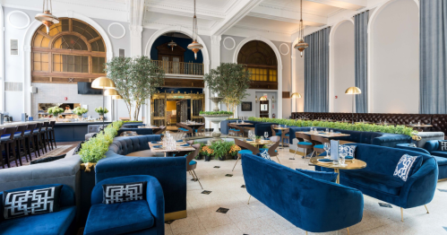 Elegant restaurant interior with blue velvet seating, greenery, and stylish decor, featuring high ceilings and warm lighting.