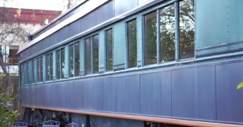 Side view of a vintage train car with multiple windows, set against a backdrop of trees and a building.