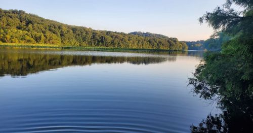 Serene river scene with calm water reflecting lush green hills under a clear sky.