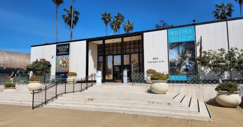 Exterior of a museum with palm trees, featuring banners for an exhibition, and a wide staircase leading to the entrance.