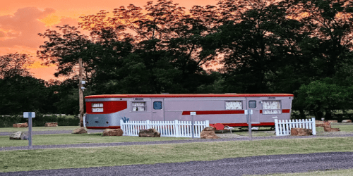 A vintage trailer with red and gray colors sits in a grassy area, framed by a white picket fence against a sunset sky.