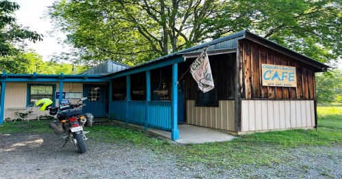 A rustic café with a blue and wooden exterior, featuring a motorcycle parked outside and trees in the background.