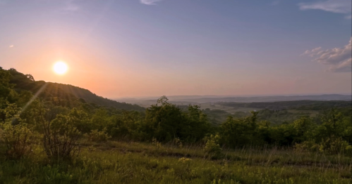 A serene landscape at sunset, with rolling hills and lush greenery under a colorful sky.
