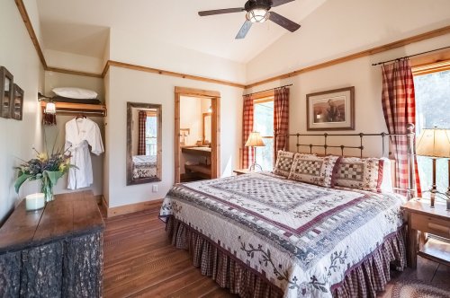 Cozy bedroom with a quilted bed, wooden accents, and natural light from windows, featuring a robe and decorative lamps.