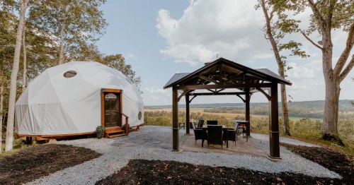 A geodesic dome cabin beside a gazebo with seating, surrounded by trees and a scenic view of rolling hills.