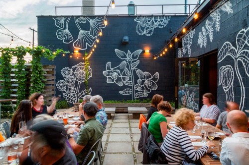 Outdoor dining scene with people at a long table, surrounded by string lights and floral wall murals.
