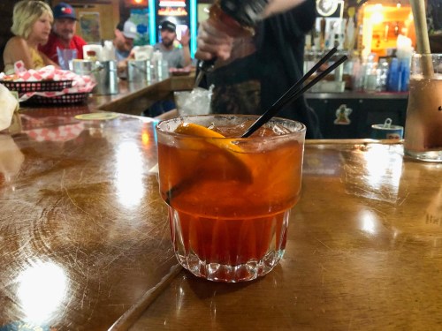 A cocktail with an orange slice sits on a wooden bar, with blurred patrons in the background.