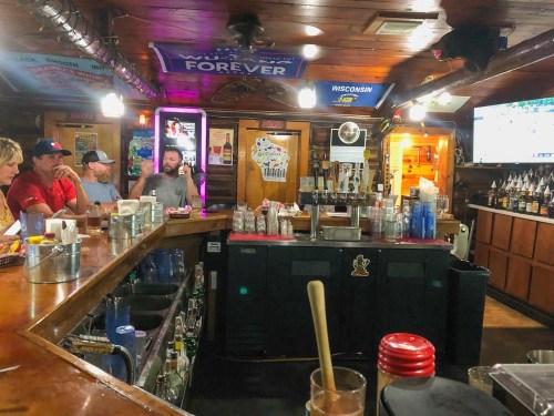 A cozy bar scene with patrons at the counter, drinks on display, and a TV showing a game in a rustic setting.