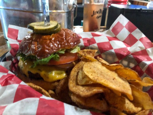 A juicy cheeseburger with lettuce, tomato, and pickles, served with crispy potato chips in a red-checkered basket.