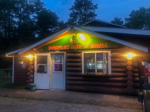 A log cabin-style building with a sign reading "Pickles Bar & Grill," illuminated at dusk with trees in the background.